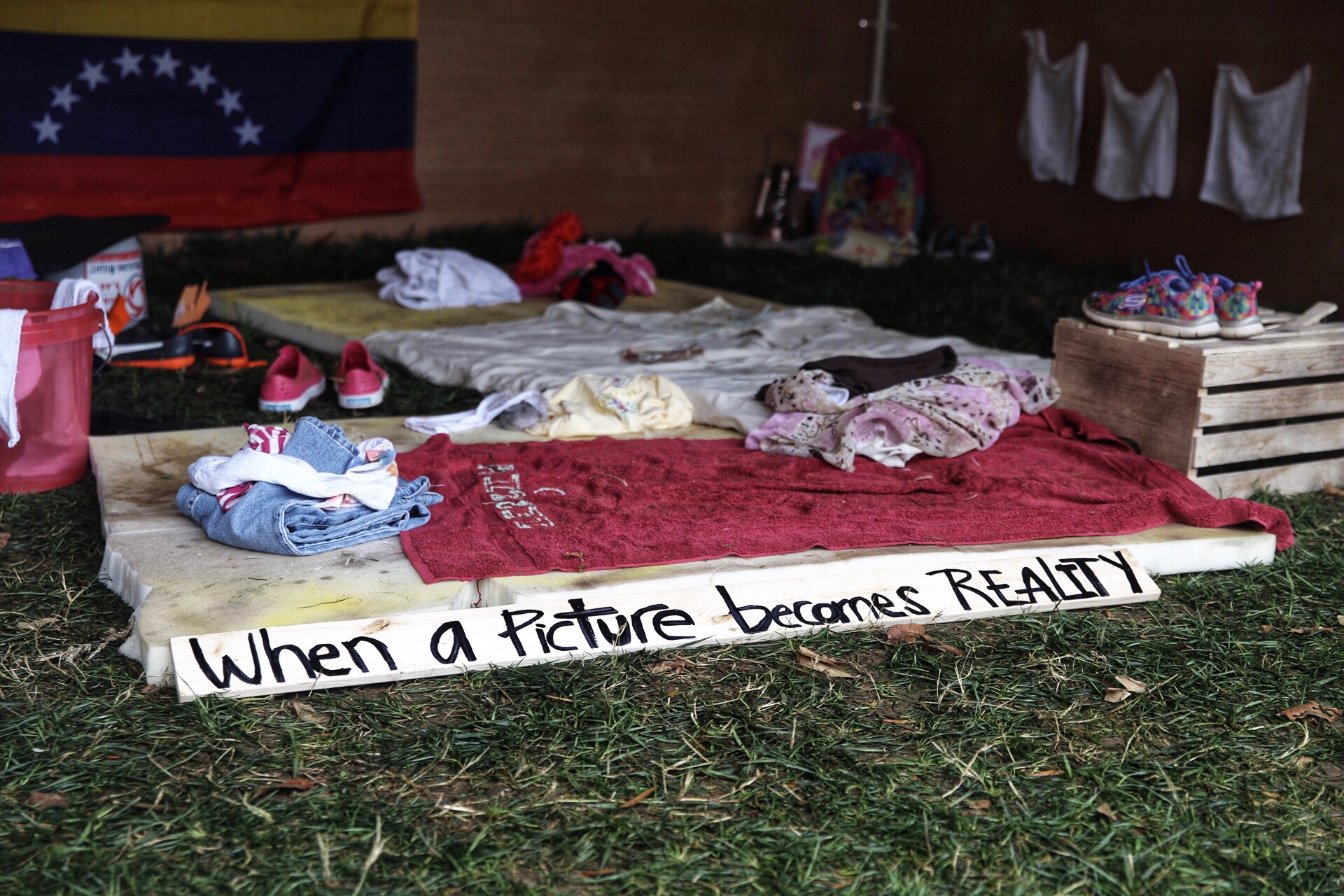 Interior of a tent showcasing the current humanitarian crisis in Venezuela with dirty mattresses, torn towels, ripped clothing, shoes with no laces, an empty water jug, and a pair of children’s stuffed animals.