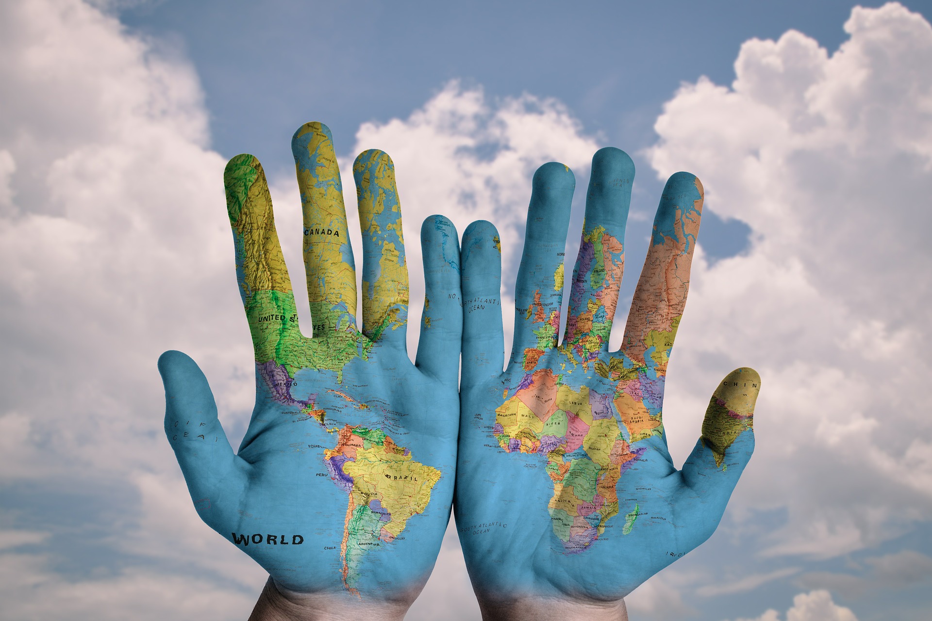 World Map painted on hands with a backdrop of puffy white clouds and blue skies.